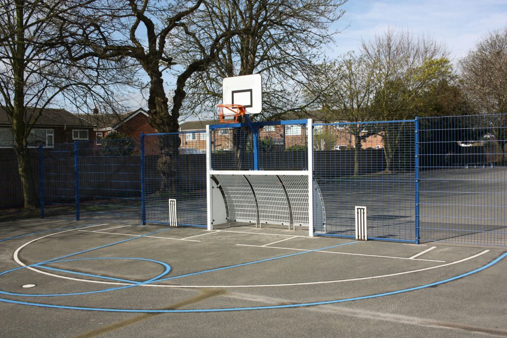 Image of a Multi Use Games Area (MUGA). This includes a white football or hockey net, a white and red basketball hoop above it, and markings in white and blue to various other sports, all surrounded by a blue fence