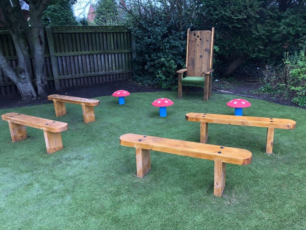 Photograph of The Playground Company’s playground project for an outdoor storytelling area for Key Stage 1 students at a school in Royal Leamington Spa. This includes a high backed wooden chair at one end and seating for students including three red, rubber topped seating mushrooms and four timber backless benches on green artificial play grass