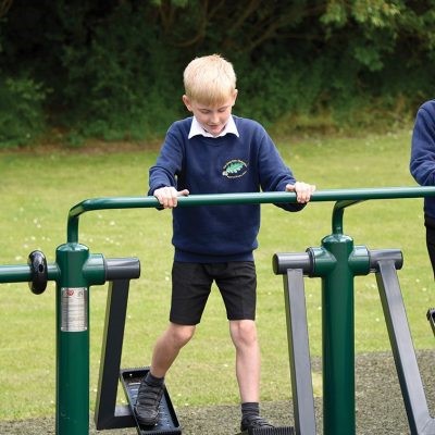 Photograph of school-children using and enjoying The School Playground Company’s product- Outdo-child-DBLHE- Double Health Walker. Standing on the black foot holds and holding onto the green safety bar, children take part in a light cardiovascular workout side by side with their peers.