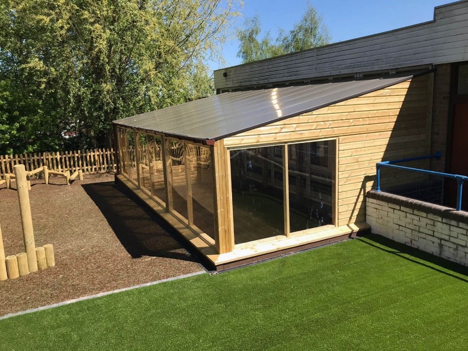 Our outdoor classroom playground shelter timber shelter in a school playground. The timber structure ajoins a larger building with floor to ceiling windows around the front and sides and a slanted Perspex roof