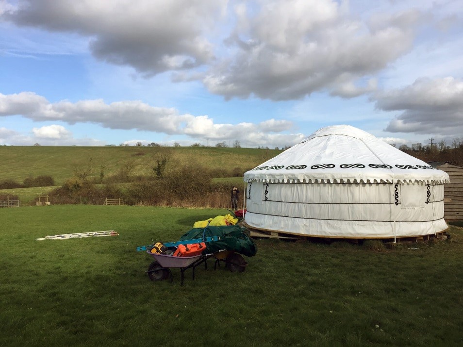 Our outdoor classroom playground shelter yurt shelters in a grass playground field. The yurt is made of white canvas material in a circular shape with a rounded triangle roof decorated with black swirling designs and embellishments.