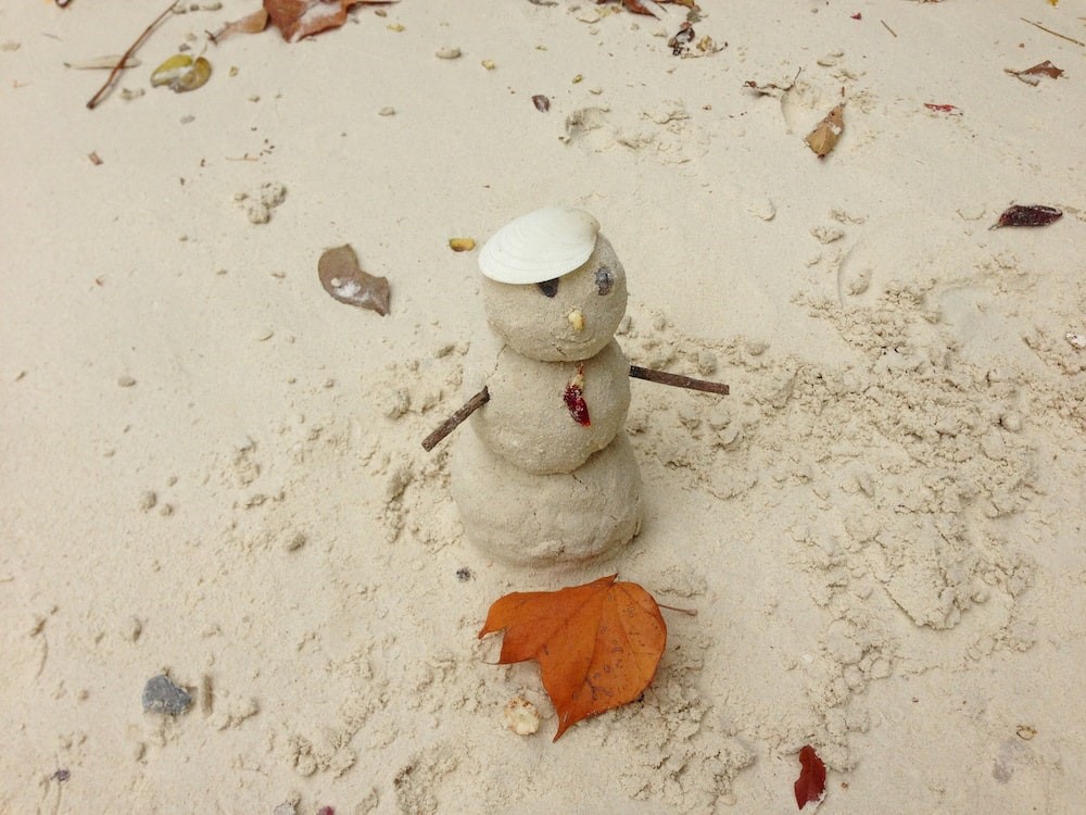 A small snowman made of sand with three round segments, two sticks for arms, small stones as buttons, eyes and nose, with a line for a smile and a white shell for a hat.