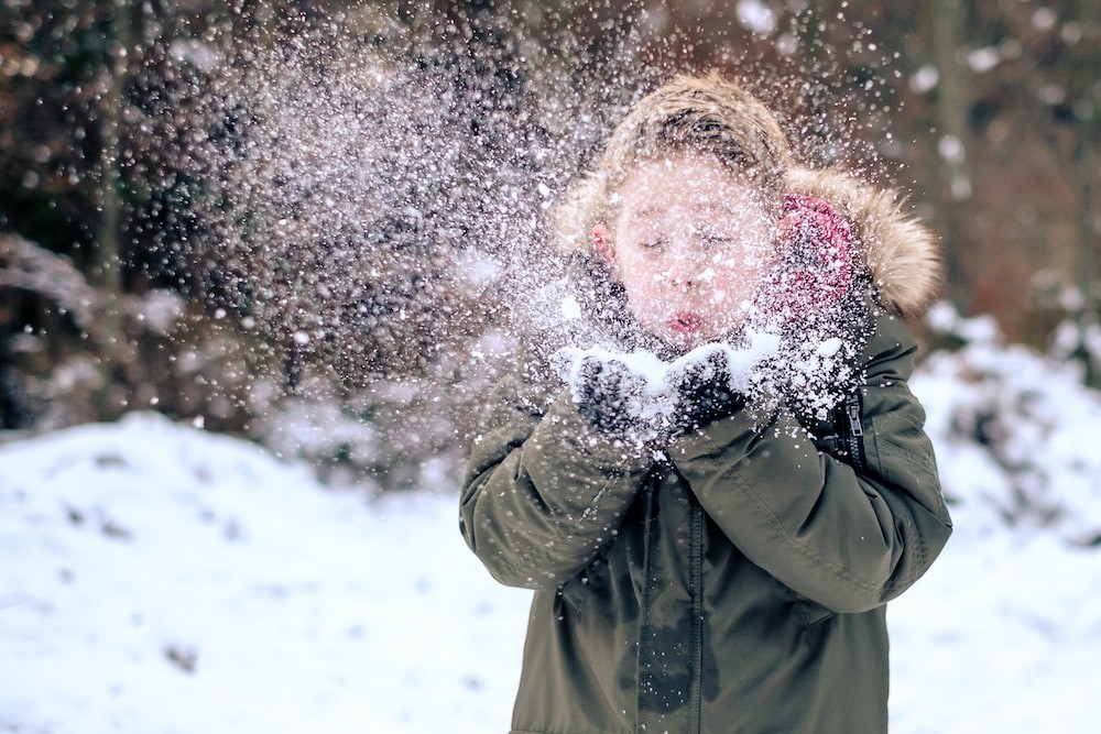 A child in a thick winter coat blows snow out of their hands at the camera.