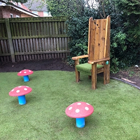 Red rubber toadstool mushroom seats around wooden high-backed storytelling chair on artificial grass playground