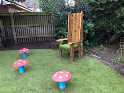 Red rubber toadstool mushroom seats around wooden high-backed storytelling chair on artificial grass playground