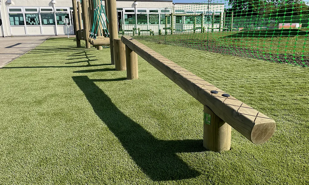 Wooden balance beam positioned low to the ground with criss-crossed markings for slip-resistance