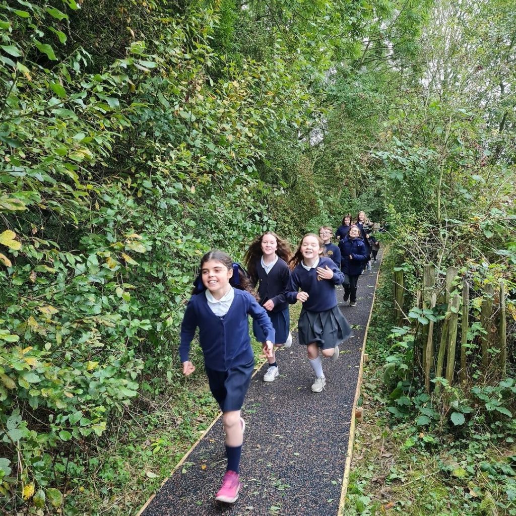 Children running along a daily mile track through a wooded area in an Oxfordshire school sports and play area